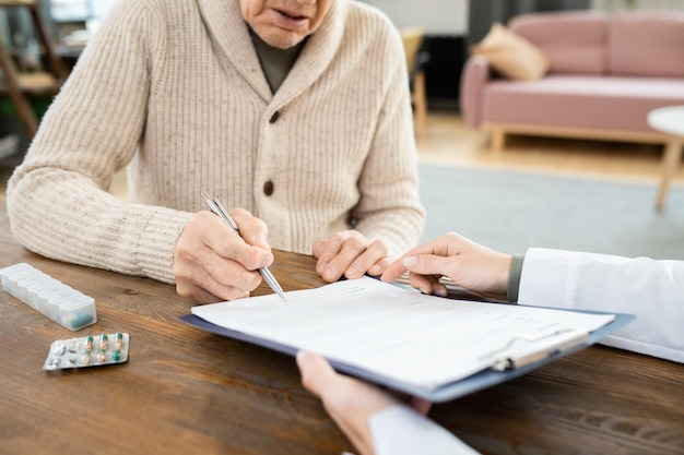 Kranker älterer Mann in Freizeitkleidung, der ein medizinisches Dokument in der Zwischenablage unterschreibt, während er am Tisch vor einer Ärztin sitzt, die auf Papier zeigt