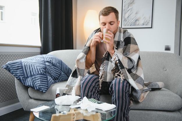 Kranker bärtiger Mann, der eine schlimme Erkältung oder saisonale Grippe hat, sitzt zu Hause auf der Couch Kerl mit Fieber, der warmes Plaid trägt und mit besorgtem Gesichtsausdruck zittert