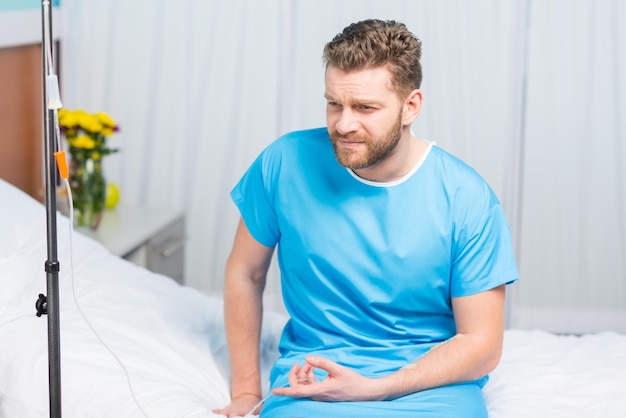 Kranker bärtiger Mann sitzt auf Krankenhausbett mit Tropfenzähler