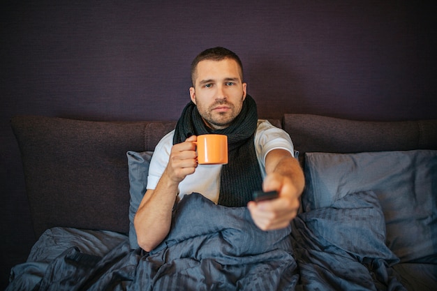 Kranker junger Mann sitzt auf Bett im Schlafzimmer und benutzt Fernbedienung. Er schaut geradeaus. Guy hat eine orangefarbene Tasse in den Händen. Er ist ruhig und konzentriert. Es gibt einen Schal um seinen Hals.