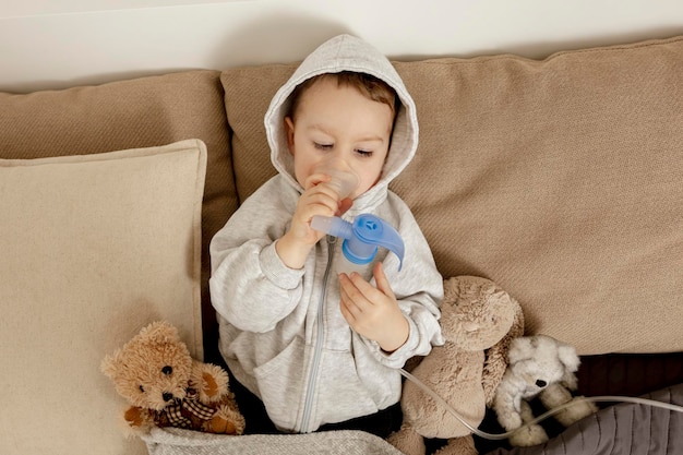 Kranker kleiner Junge mit Inhalator zur Hustenbehandlung. Unwohles Kind, das auf seinem Bett inhaliert. Grippe-Saison. Medizinisches Verfahren zu Hause. Interieur und Kleidung in natürlichen Erdfarben. Gemütliche Umgebung.