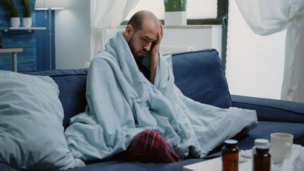 Kranker Mann reibt Schläfen gegen Kopfschmerzen und Erkältung in Decke gehüllt. Erwachsener mit Krankheit und Migräne mit Virussymptomen und Medikamenten auf dem Tisch, um Krankheiten zu heilen. Person mit Grippe