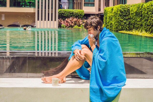 Kranker Mann Reisender. Der Mann hat sich im Urlaub erkältet, sitzt traurig am Pool, trinkt Tee und putzt sich die Nase in eine Serviette.