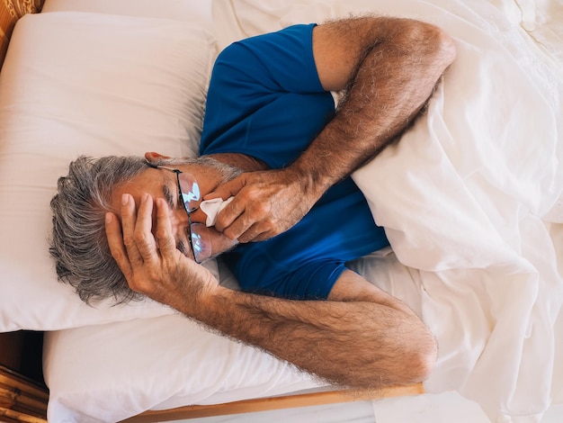 Kranker reifer Mann, der im Bett liegt und sich mit einem Taschentuch die Nase weht