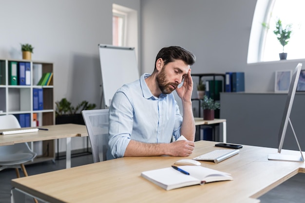 Kranker und müder junger Mann bei der Arbeit Büroangestellter-Manager-Freiberufler, der an einem Schreibtisch im Büro sitzt, fühlt sich nicht wohl, wischt sich die Nase ab, hat eine laufende Nase, hustet, hält den Kopf