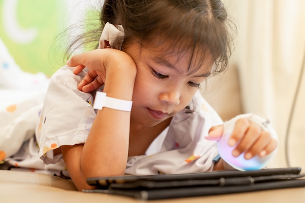 Krankes asiatisches kleines Kindermädchen, das IV Lösung verbunden hat, digitale Tablette spielend