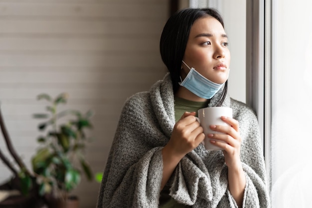 Krankes asiatisches Mädchen in medizinischer Gesichtsmaske, das am Fenster steht und sich danach sehnt, nach draußen zu gehen, während es auf Quar...