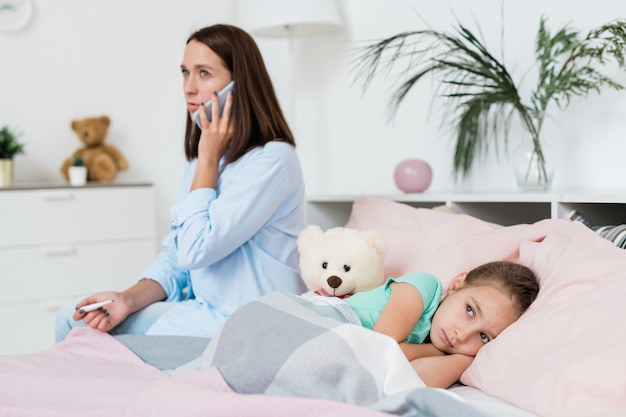 Krankes kleines Mädchen, das auf Bett unter Decke liegt, während ihre Mutter Arzt auf Handy ruft