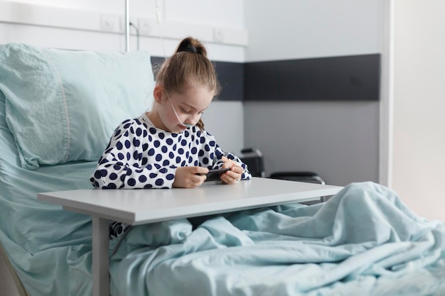 Krankes kleines Mädchen, das auf dem Smartphone spielt, während es sich auf dem Patientenbett im Zimmer der Kinderklinik ausruht. Kranker junger Patient, der Handyunterhaltung genießt, während er auf der Genesungsstation des Krankenhauses sitzt.