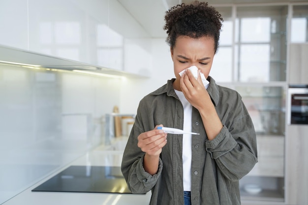 Krankes Mischlingsmädchen, das die Nase bläst, überprüft die Temperatur mit einem elektronischen Thermometer zu Hause Grippefieber