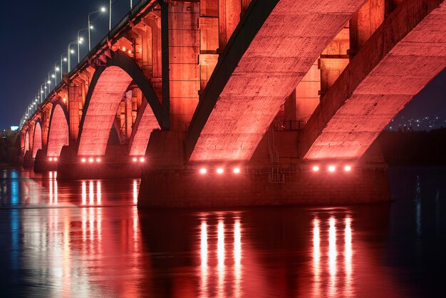 Foto krasnoyarsk, rusia. vista nocturna del puente comunal de krasnoyarsk con luz de fondo roja y el río yenisey.