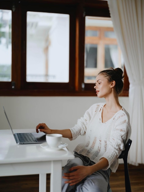 Kreative junge Frau, die zu Hause an einem Schreibtisch arbeitet, in einem gemütlichen Interieur mit einer Tasse Getränk, Home-Office-Freelancer mit einem Lächeln, Kommunikation und Online-Ausbildung