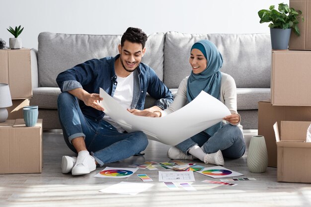 Kreative junge muslimische Familie, die an Design für ihr neues Haus arbeitet, auf dem Boden zwischen Kisten mit Habseligkeiten sitzt, Wohnungsplan und Farbpalette hält, über Wohndesign nachdenkt, Raum kopieren