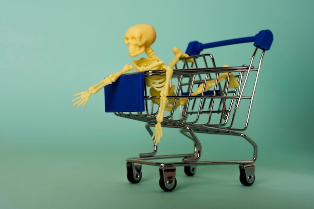 Foto kreative minimalistische komposition, ein skelett, das auf einem supermarktwagen reitet, türkisfarbener hintergrund, ein schädel.