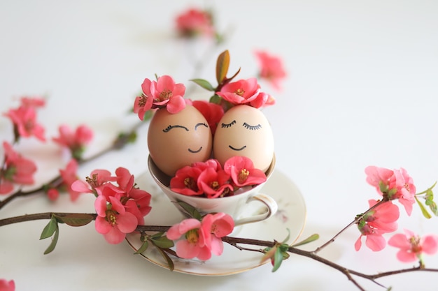 Kreative Ostereier mit süßen, schläfrigen Augen und Frühlingsblumen in Teetasse auf weißem Hintergrund