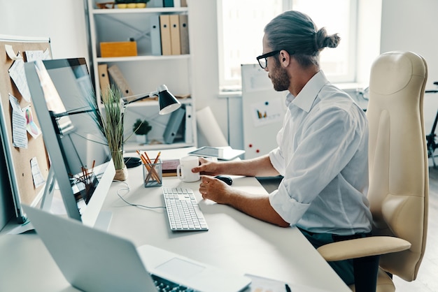 Kreativer Geschäftsmann. Nachdenklicher junger Mann im Hemd, der mit Computer arbeitet und Kaffee trinkt, während er im Büro sitzt