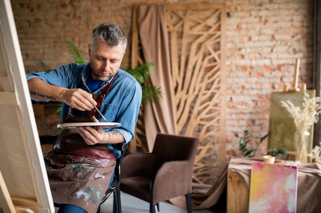 Kreativer Mann mittleren Alters in der Schürze, die Farben auf Palette mischt, bevor über neue Malerei im Studio gearbeitet wird