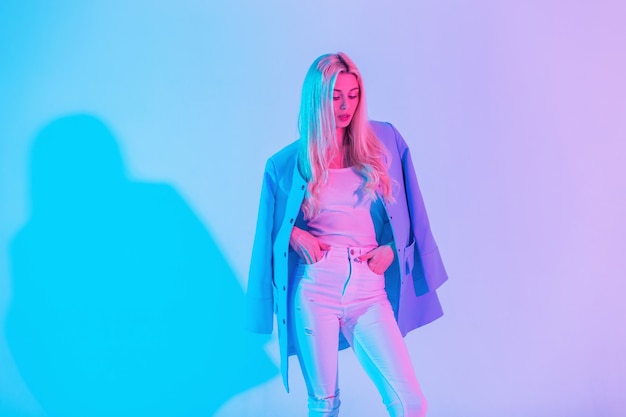 Kreatives buntes Porträt eines jungen schönen Mädchens in modischer eleganter Kleidung mit blauem Mantel, weißer Bluse und Jeans im Studio vor einem Hintergrund von neonrosa Licht