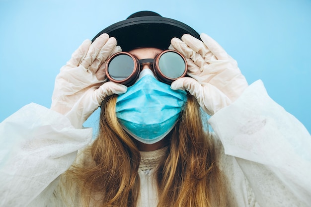 Kreatives Mädchen in einem weißen Gewand, einer medizinischen Maske, einer Brille und einem Hut auf einem blauen Hintergrund. Selbstisolation während der Coronavirus-Quarantäne. Epidemic covid-2019.