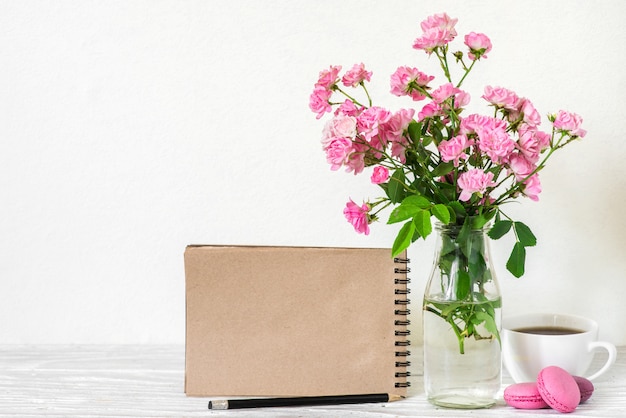 Kreatives Modell mit einem schönen Blumenstrauß aus rosa Rosenblumen, Kaffeetasse, Makronen und leerem Hipster-Notizbuch