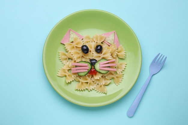 Kreatives Servieren für Kinder Teller mit süßer Katze aus leckerem Nudelgemüse und Wurst auf hellblauem Hintergrund flach liegend