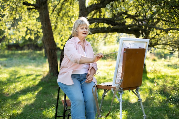 Kreativität, Hobby, Kunst, Freizeit und Menschenkonzept. Porträt einer glücklichen älteren Künstlerin mit Pinsel, die im Sommer auf Staffelei im Freien im Freien im grünen Park zeichnet.