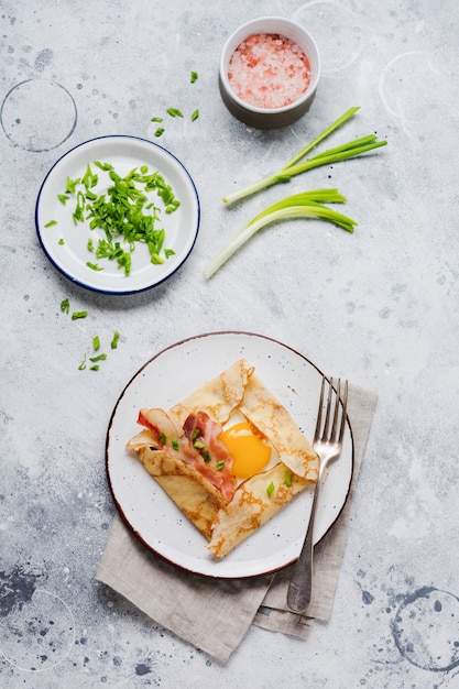 Krepp mit Spiegelei, Käse, Speck und Frühlingszwiebeln zum Frühstück auf hellgrauer Betonoberfläche