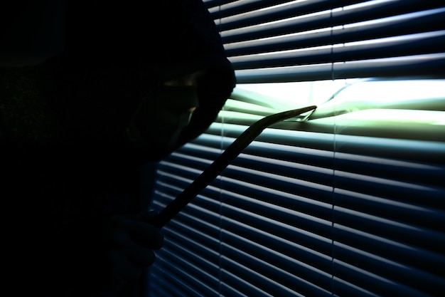 Krimineller mit Brecheisen spioniert durch das Fenster. Alte Rollladenjalousien.