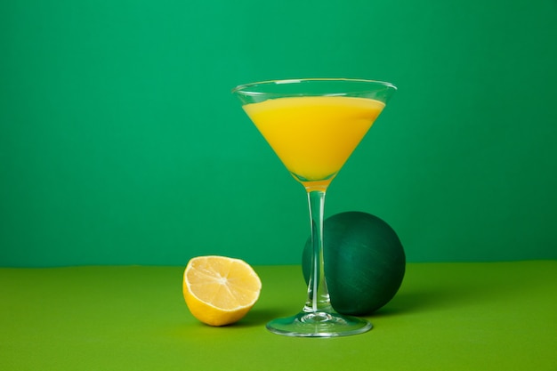Kristallglas mit frischem Alkoholgetränk serviert auf dem Tisch in der Nähe von saftiger halbierter Zitrone und dekorativer Kugel vor grünem Hintergrund