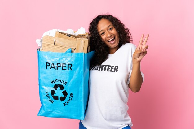 Kubanisches Mädchen des Teenagers, das eine Recycling-Tasche voll Papier hält, um lokalisiert auf rosa lächelnd und siegreiches Zeichen zu recyceln