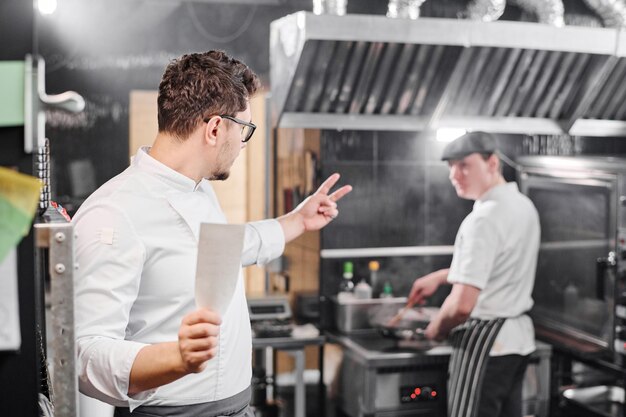Küchenchef gestikuliert seinem Assistenten, Essen für zwei Personen während ihrer Arbeit in der Großküche zuzubereiten