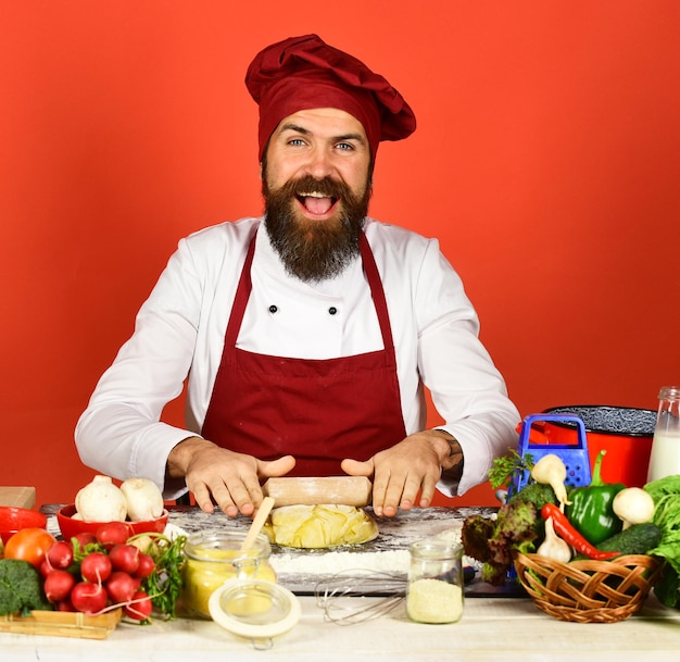 Küchenchef mit Gewürzen, Gemüse und Teig auf dem Tisch Mann mit Bart sitzt an der Arbeitsplatte auf rotem Hintergrund Koch mit fröhlichem Gesicht in burgunderfarbener Uniform rollt Teig mit Nudelholz Konzept des Kochvorgangs
