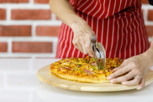 Küchenchefin in roter Streifenschürze mit Rollklinge, um köstliche frisch gebackene Pizza auf Holzplatte in der Küche sorgfältig aufzuteilen, um traditionelles italienisches Essen leicht zu essen