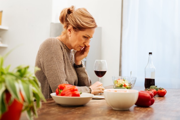 Küchentisch. Beunruhigte hellhaarige Frau, die mit ihrem Lebensstil depressiv ist, während sie allein Alkohol trinkt