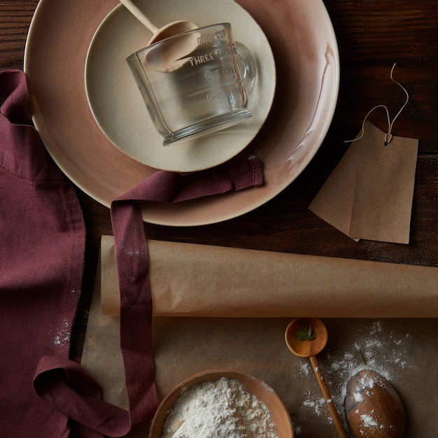 Küchenzubehör mit Teller, Backpapier und Schürze auf Holzuntergrund mit Mehl in einer Schüssel zum Backen von Kuchen oder Pfannkuchen, flach