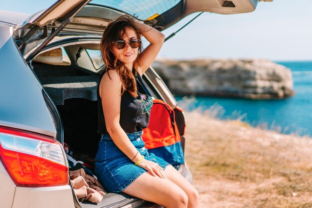 Kühle junge Frau in der Sonnenbrille sitzt auf dem Kofferraum eines Autos mit Meerblick, Autofahrkonzept