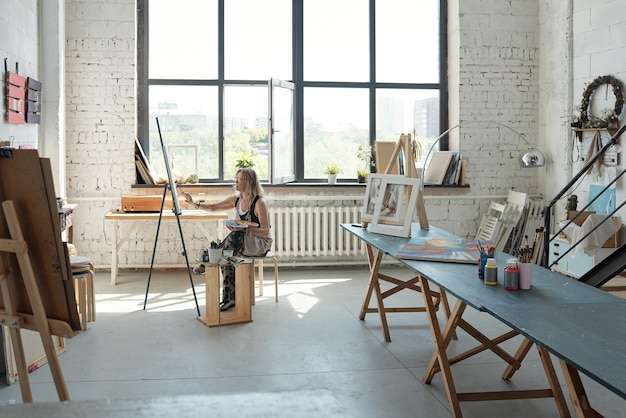 Künstlerin sitzt vorne auf Leinwand und malt Bild in eigenem geräumigen Studio