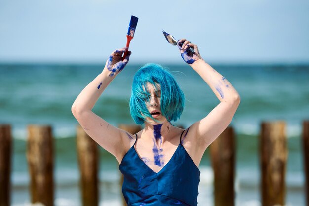 Künstlerische blauhaarige Frau Performance-Künstlerin in dunkelblauem Kleid mit blauen Gouache-Farben beschmiert Malerei und Tanz am Strand. Bodypainting- und Body-Art-Konzept, ausdrucksstarke Performance-Kunst im Freien