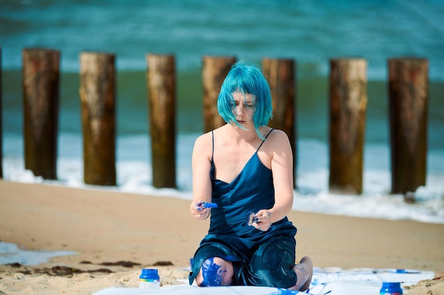 Künstlerische blauhaarige Frau Performance-Künstlerin in dunkelblauem Kleid mit blauer Gouache-Malerei beschmiert, während sie am Strand sitzt. Bodypainting- und Body-Art-Konzept, ausdrucksstarke Performance-Kunst im Freien