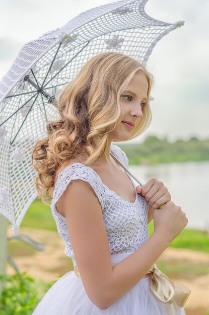 Künstlerisches leichtes Sommerporträt blondes Mädchen in einem weißen Kleid am Flussufer im Vintage-Stil