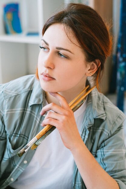 Künstlerporträt Kreativer Lebensstil Fantasietalent Inspirierte friedliche, nachdenkliche Frau mit Pinseln, die im Lichtkunststudio tagträumen