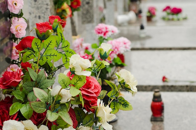 Künstliche Blumen auf dem Grab auf dem Friedhof mit brennender roter Kerze