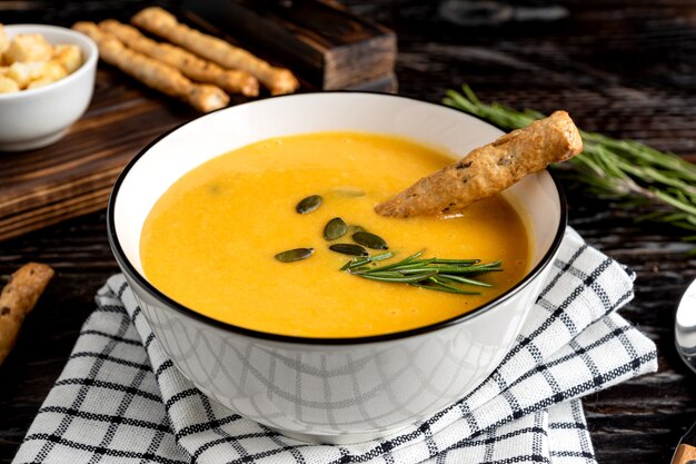 Kürbis-Karotten-Suppe mit Sahne, Kürbiskernen, Croutons und Grossini-Brotstangen auf dar k Holzhintergrund. Rustikaler Stil, selektiver Fokus.