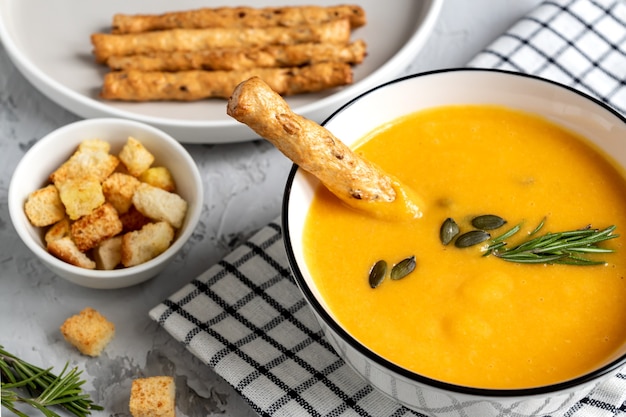 Kürbis-karotten-suppe mit sahne, kürbiskernen, croutons und grossini-brotstangen auf dar k holzhintergrund. rustikaler stil, selektiver fokus.