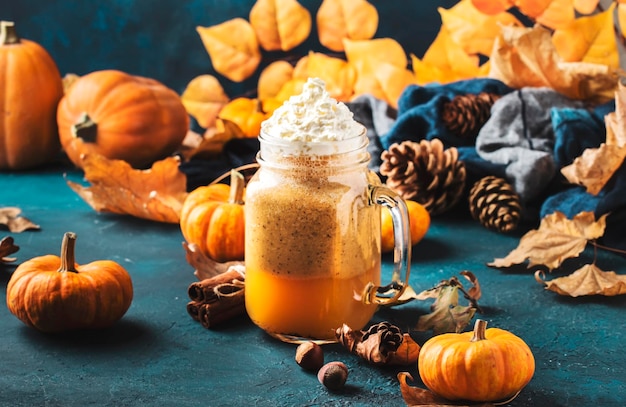 Kürbisgewürzter Latte oder Kaffee im Glasgefäß auf blauem Tisch Herbst- oder Winter-Heißgetränk in festlicher natürlicher Tischdekoration mit Orangenblättern würzt kleine Pumkins-Tannenzapfen