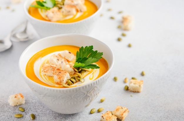 Kürbissuppe mit Sahne, Croutons, Kürbiskernen und Petersilie auf einem grauen Beton- oder Steinhintergrund.