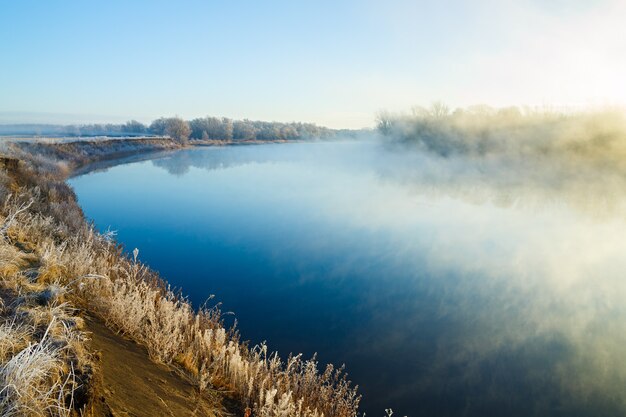 Küste mit frostbedecktem Gras. Fluss mit Morgennebel.