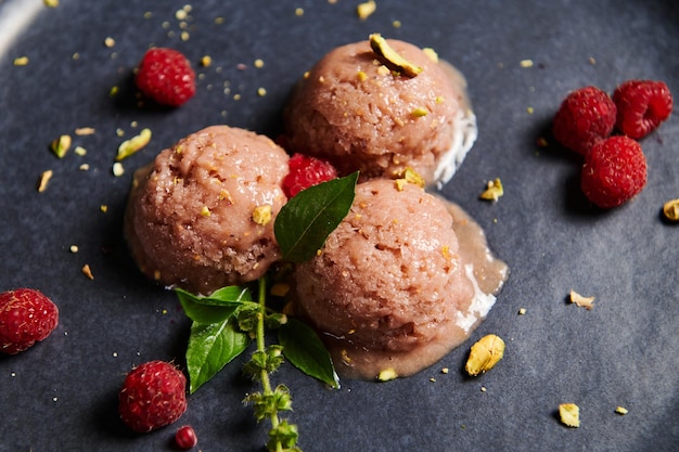 Kugeln Himbeersorbet mit bestreuten grünen Pistazien, Himbeeren und Zitronenbasilikum auf dunkelblauem Teller Nahaufnahme