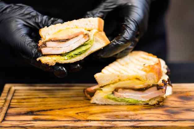 Kulinarische Meisterklasse. Nahaufnahme der Chefhände, die eine Hälfte des geräucherten Truthahnbrustsandwiches über Holzbrett halten.