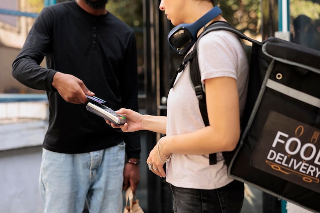 Kunde, der kontaktlose Zahlung für die Lieferung von Lebensmitteln mit Kreditkarte in Nahaufnahme leistet. Kurier mit Rucksack, der ein POS-Terminal hält, afroamerikanischer Kunde, der für das Essen zum Mitnehmen im Restaurant bezahlt, Nahaufnahme